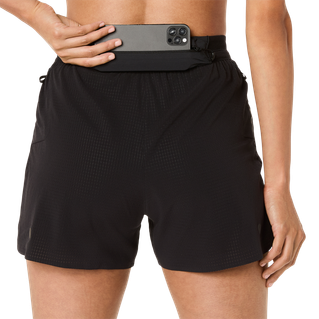 En person set bagfra er iført ASICS Metarun Split Shorts, Dame, Performance Black, og holder en sort smartphone delvist gemt i linningen mod en almindelig hvid baggrund.