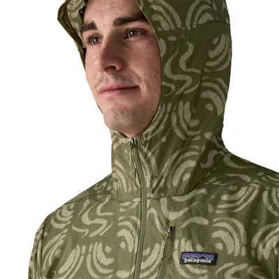 En person er iført en sandbølger/grøn Patagonia Houdini Jacket, herre, med lynlås i nakken. Den lette vind- og regntætte jakke har det karakteristiske Patagonia-logo på brystet. Baggrunden er hvid.