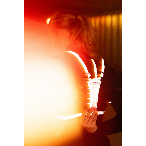 En person med tilbagestrøget blondt hår ses bagfra iført BeeSafe LED Harness USB Phone Pocket, Unisex, Silver, og med en enhed i hånden. Kraftigt orange lys dækker delvist forgrunden; baggrunden er svagt oplyst.
