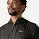 En mand med kort krøllet hår og skæg bærer Patagonia Houdini Jacket, Herre, Black w/Black, logoet er synligt på venstre bryst. Han kigger lidt opad og til venstre mod en ensfarvet baggrund i denne lette vind- og regnjakke.