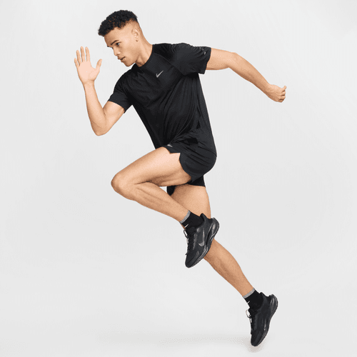 En mand i en sort Nike Stride SS Running Top, Herre, Black/Reflective Silv og shorts med sneakers er fanget midt i et spring i en løbepositur mod en almindelig, lys baggrund, der fremhæver topens præstationsegenskaber.