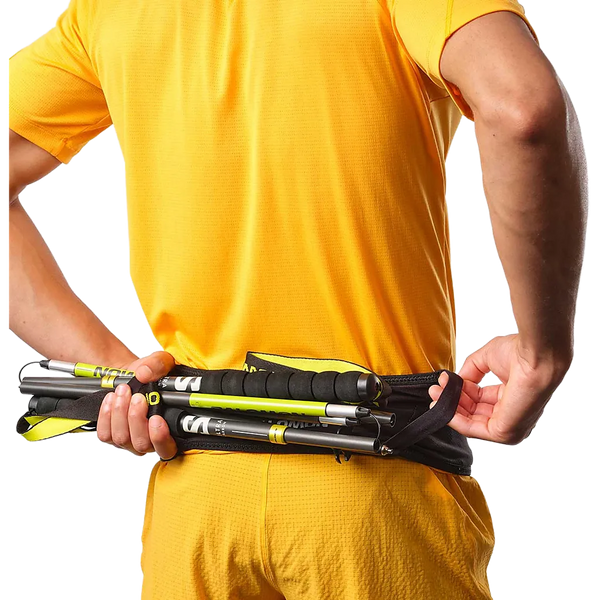 En person i gult holder en foldet vandrestav og et sort Salomon Pulse Løbebælte, Unisex, med den ene hånd trukket i bæltets lynlås bag ryggen.