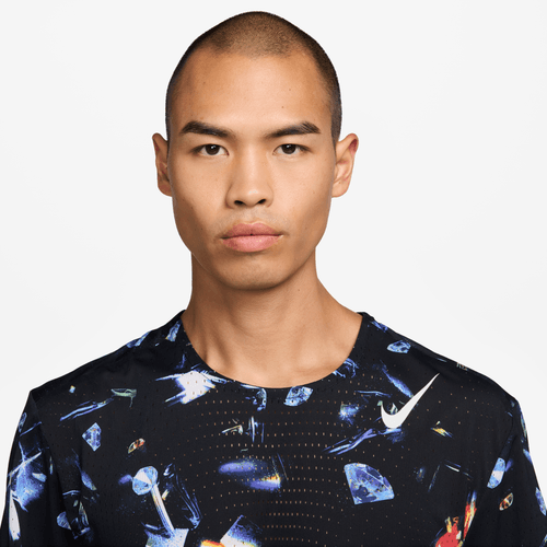 En person med barberet hoved bærer Nike Aeroswift Aop Ss Running T-Shirt til mænd i Black/Laser Orange med et farverigt, abstrakt mønster og ser frem for sig med et neutralt udtryk mod en almindelig hvid baggrund.