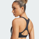 En kvinde med krøllet hår i en knold bærer Adidas Adizero Medium Support Run Bra, Dame, Black - med hvide striber på den ene side. Hun kigger sig over skulderen, klar til løb, mod en almindelig lys baggrund.