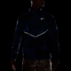 En person iført en mørk Nike Miler UV Repel-løbejakke til mænd i farven Midnight Navy med reflekterende sølvdetaljer og mønstrede shorts står foran en sort baggrund. Nike-logoet og de reflekterende striber på jakken lyser op.