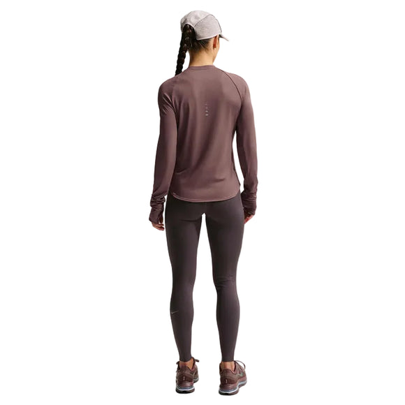En person står med ryggen til og har en lilla Nike Swift UV langærmet løbetrøje til kvinder i farven »Tattoo/Reflective Silver« på sammen med matchende leggings, løbesko og en lys kasket. Håret er sat op i to fletninger foran en ensfarvet, lys baggrund. Dette sæt fra Nike kombinerer stil og UV-beskyttelse til aktive dage udendørs.