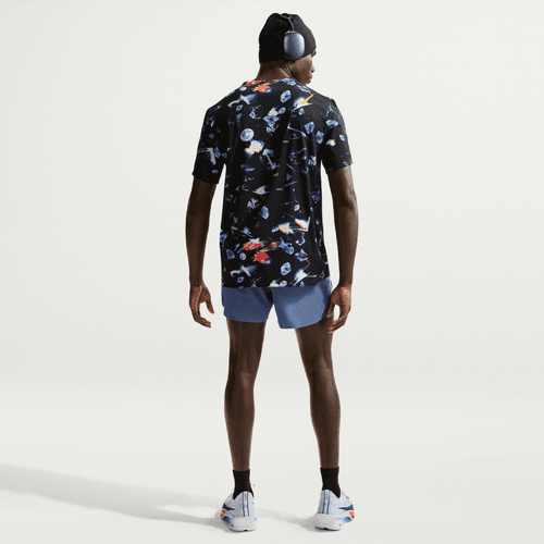 En person står med ryggen til og er iført en Nike Aeroswift Aop Ss Running T-Shirt til mænd i sort/laser orange med et farverigt print, lyseblå shorts, hvide løbesko, en sort hue og hovedtelefoner. Baggrunden er helt hvid.