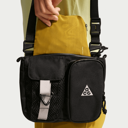 En person lægger en gul pose i en sort Nike ACG Vest (Dame, Peat Moss/Lt Liquid Lime/Summit White) crossbody-taske med flere rum, netlomme, spændelukning og et ACG-logo på forsiden mod en lys baggrund.