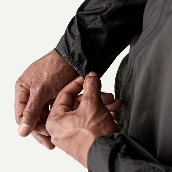 Nærbillede af en persons hænder, der justerer den elastiske manchet på Patagonia Houdini Jacket, Herre, Black w/Black mod en almindelig lys baggrund. Kun hænderne, en del af armene og jakken er synlige.