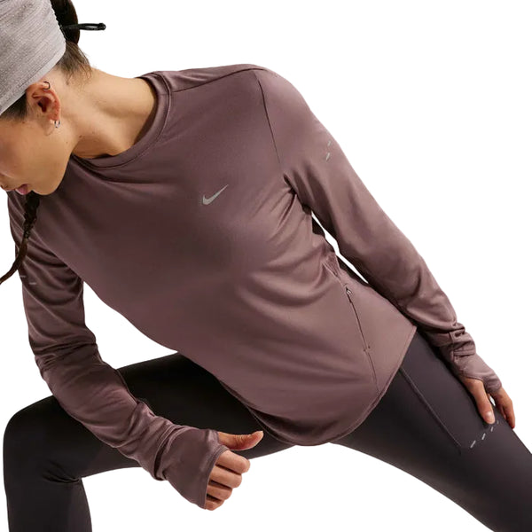 En kvinde i en lilla Nike Swift UV Long Sleeve Shirt, Dame, Tattoo/Reflective Silv og sorte leggings strækker sig frem med den ene arm strakt frem og den anden bøjet. Hendes hoved er vendt nedad, delvist dækket af et lysegråt pandebånd. UV-beskyttelse beskytter hende udendørs.