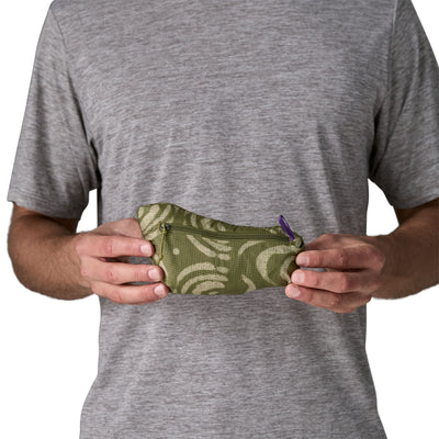 En person i grå t-shirt holder en lille, grøn lynlåspose med et let hvirvelmønster og lilla lynlåstræk - ideel til vigtige ting som Patagonia Houdini Jacket, Herre, Sand Waves/Caper Green. Pungen holdes i brysthøjde med begge hænder.