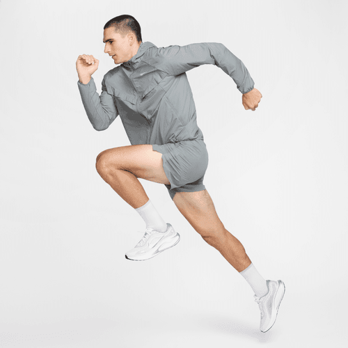 En mand i Nike Repel UV Running Jacket Herre (Smoke Grey/Reflective Silv), matchende shorts, hvide sokker og sneakers springer i luften mod en hvid baggrund. Jakken giver UV-beskyttelse til udendørs løbeture.