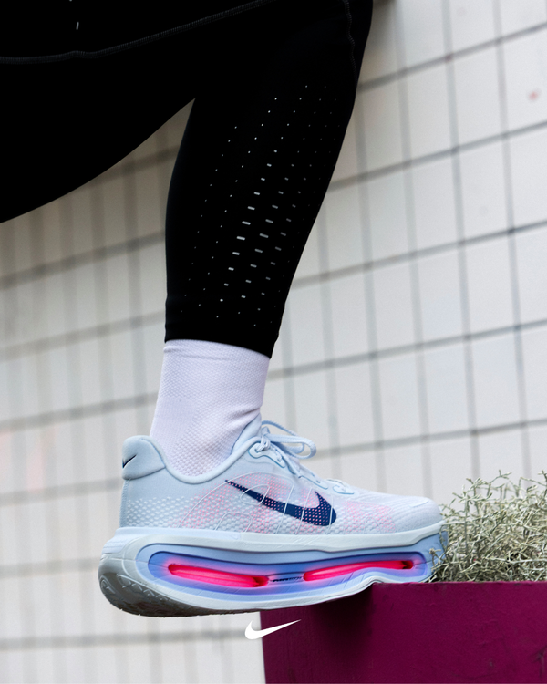 En person iført hvide Nike-sneakers med blå swoosh, hvide sokker og sorte leggings træder ned på et lyserødt underlag foran en flisebelagt væg. Sneakersne har synlig pink støddæmpning i sålen.