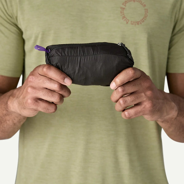 En person i en lysegrøn t-shirt holder en lille, sort Patagonia-pose med en lilla trækfane - ideel til opbevaring af vigtige ting som Houdini Jacket, Herre, Black w/Black - sikkert med begge hænder mod en ensfarvet baggrund.