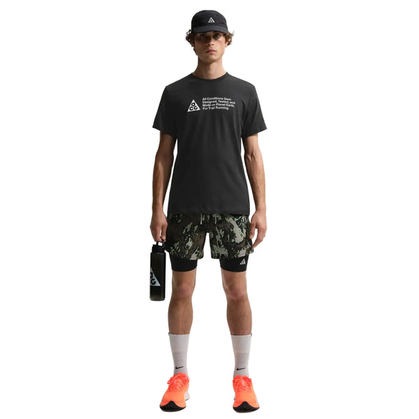 En person ser fremad mod en almindelig hvid baggrund, iført en sort Nike ACG Df Tee Std Trail Logo Herre t-shirt, sort kasket, camouflageshorts, hvide sokker, neonorange sneakers og med en sort vandflaske i hånden.
