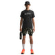En person ser fremad mod en almindelig hvid baggrund, iført en sort Nike ACG Df Tee Std Trail Logo Herre t-shirt, sort kasket, camouflageshorts, hvide sokker, neonorange sneakers og med en sort vandflaske i hånden.