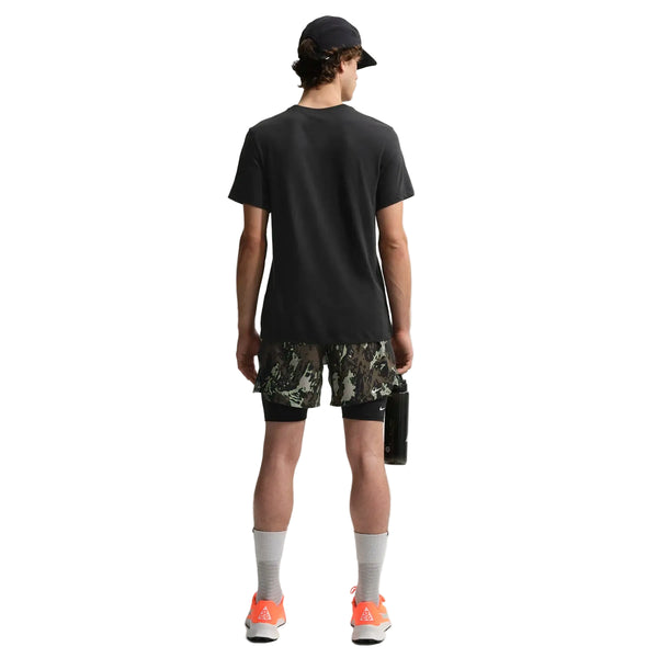 En person står med ryggen til og er iført Nike ACG Df Tee Std Trail Logo t-shirt i sort, camouflageshorts, grå sokker og orange sneakers, mens han holder en rektangulær genstand i venstre hånd mod en hvid baggrund.