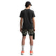 En person står med ryggen til og er iført Nike ACG Df Tee Std Trail Logo t-shirt i sort, camouflageshorts, grå sokker og orange sneakers, mens han holder en rektangulær genstand i venstre hånd mod en hvid baggrund.