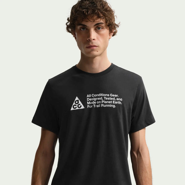 En person med krøllet hår er iført en sort Nike ACG Df Tee Std Trail Logo, Herre t-shirt med hvidt logo og tekst på brystet: "All Conditions Gear. Designet, testet og fremstillet på planeten Jorden. Til trailløb.