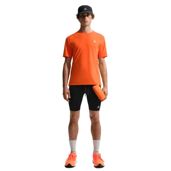 En person ser fremad i en Nike ACG Dfadv Solar Chase Ss tee (Safety Orange/Summit White), sorte shorts, hvide sokker, orange sko og en sort kasket med en orange vandflaske i hånden. Baggrunden er almindelig hvid.