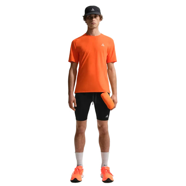 En person ser fremad i en Nike ACG Dfadv Solar Chase Ss tee (Safety Orange/Summit White), sorte shorts, hvide sokker, orange sko og en sort kasket med en orange vandflaske i hånden. Baggrunden er almindelig hvid.