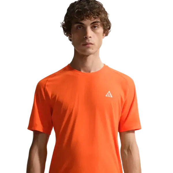 En person med kort krøllet hår bærer en Nike ACG Dfadv Solar Chase Ss træningstop i Safety Orange/Summit White med et lille hvidt trekantlogo på brystet, fremstillet af svedtransporterende materiale, stående mod en hvid baggrund.
