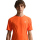 En person med kort krøllet hår bærer en Nike ACG Dfadv Solar Chase Ss træningstop i Safety Orange/Summit White med et lille hvidt trekantlogo på brystet, fremstillet af svedtransporterende materiale, stående mod en hvid baggrund.