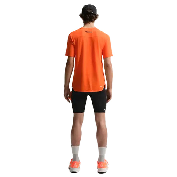 En mand kigger væk fra kameraet iført Nike ACG Dfadv Solar Chase Ss træningstop i Safety Orange/Summit White, parret med sorte shorts, hvide sokker, orange sneakers og en sort kasket. Baggrunden er helt hvid.
