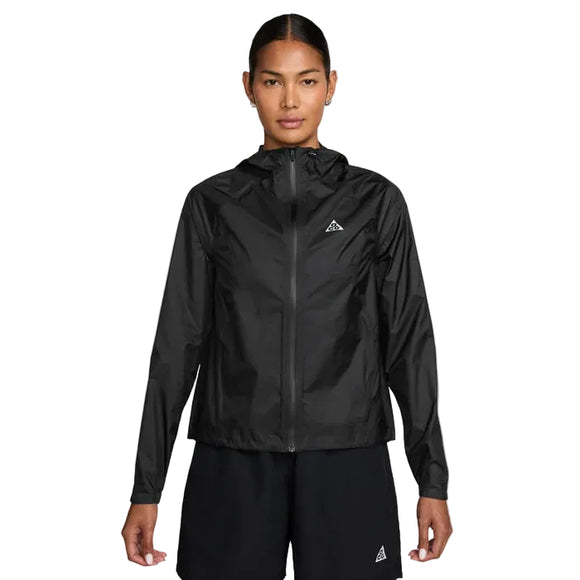 En kvinde står med ansigtet fremad i en sort Nike ACG Sfadv Trailwind Jkt, Dame, Black/Summit White - en vindtæt, vandafvisende jakke med et lille trekantet logo på venstre bryst - sammen med sorte shorts på en almindelig hvid baggrund.