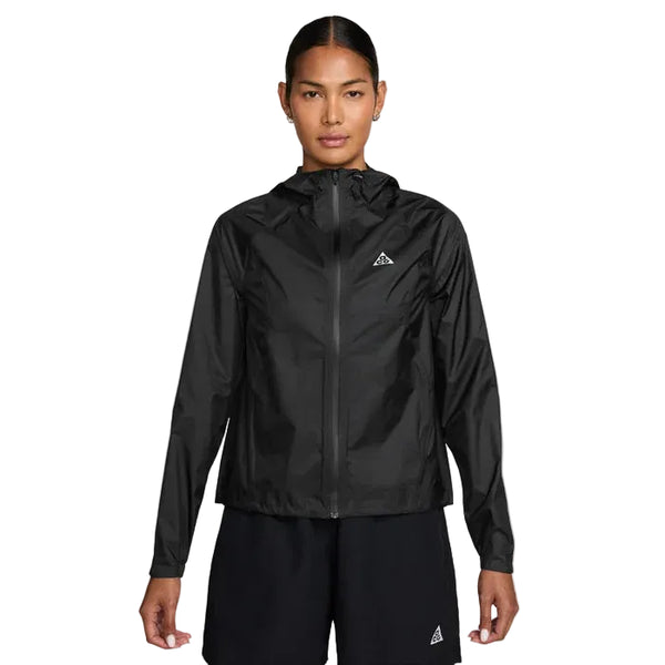 En kvinde står med ansigtet fremad i en sort Nike ACG Sfadv Trailwind Jkt, Dame, Black/Summit White - en vindtæt, vandafvisende jakke med et lille trekantet logo på venstre bryst - sammen med sorte shorts på en almindelig hvid baggrund.