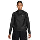 En kvinde står med ansigtet fremad i en sort Nike ACG Sfadv Trailwind Jkt, Dame, Black/Summit White - en vindtæt, vandafvisende jakke med et lille trekantet logo på venstre bryst - sammen med sorte shorts på en almindelig hvid baggrund.