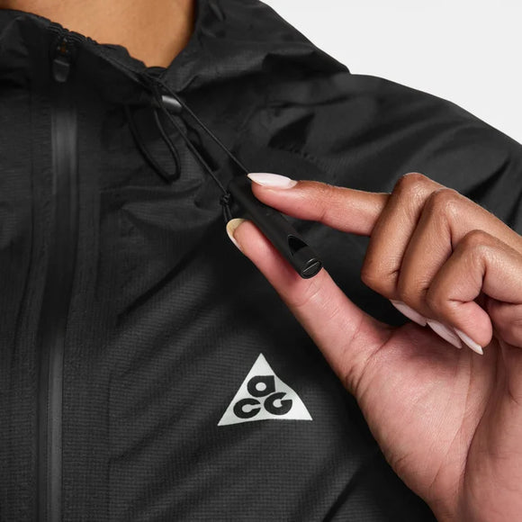 En person i en Nike ACG Sfadv Trailwind Jkt, Dame, Black/Summit White, med et hvidt ACG-trekantslogo, holder jakkens snoretræk ved kraven og fremhæver denne vindtætte vandafvisende jakke.