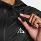 En person i en Nike ACG Sfadv Trailwind Jkt, Dame, Black/Summit White, med et hvidt ACG-trekantslogo, holder jakkens snoretræk ved kraven og fremhæver denne vindtætte vandafvisende jakke.