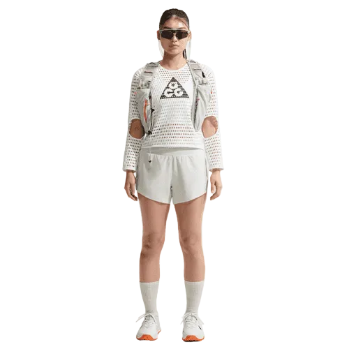 En person står med ansigtet vendt fremad iført solbriller, en Nike ACG Radical AirFlow NXT langærmet løbetrøje til kvinder i hvid/antracit, hvide shorts, en lys vest, hvide sokker og Nike-løbesko med orange såler foran en ensfarvet hvid baggrund.