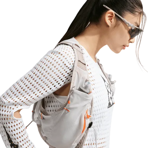 En kvinde med langt, mørkt hår sat op i en hestehale og solbriller har en hvid/antracitfarvet Nike ACG Radical AirFlow NXT langærmet løbetrøje på samt en lysegrå funktionsvest med flere lommer; hun ses i profil foran en hvid baggrund.