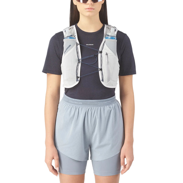 En person iført sort t-shirt, lyseblå shorts, solbriller og Salomon ADV Skin 5 væskevest (Unisex, Alloy/Gray Violet/Spellbo) med vandflasker i forlommerne står klar til terrænløb mod en almindelig hvid baggrund.