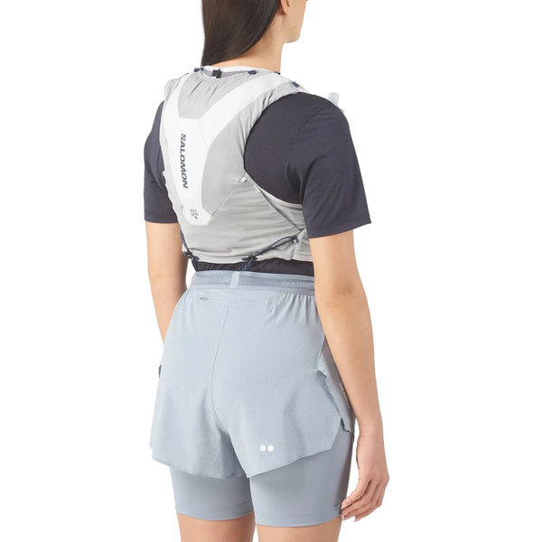En person med langt mørkt hår, i sort skjorte, lyseblå løbeshorts og en Salomon ADV Skin 5 Unisex vest i Alloy/Gray Violet/Spellbo, står med ryggen til kameraet - klar til terrænløb.