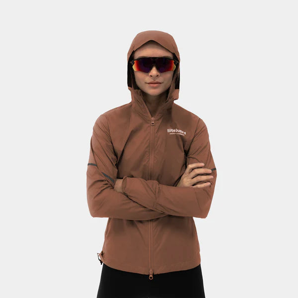 En kvinde i en kakaobrun Liiteguard Aero-Tech Windbreaker med hætten oppe og armene over kors bærer store solbriller på en lysegrå baggrund. Den åndbare damejakke har et lille Liiteguard-logo på brystet.