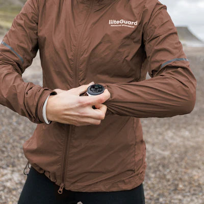 En kvinde i en kakaobrun Liiteguard Aero-Tech Windbreaker står udendørs og justerer sin manchet for at afsløre en sort håndledsanordning. Den åndbare damejakke er vand- og vindafvisende, perfekt til det barske, stenede landskab.