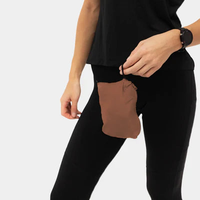 En person i Liiteguards Aero-Tech Windbreaker, Dame, Cocoa Brown holder en lille pose i bukselinningen. Den venstre hånd hviler på tasken, og et sort ur bæres på højre håndled.