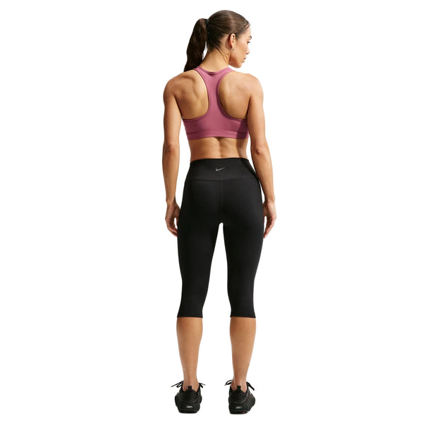 En kvinde står med ryggen til og har en Nike-sport-BH med polstring til kvinder i farven Pink Smoke/White på. Hun har desuden sorte Nike-capri-leggings med det ikoniske Swoosh-logo og Dri-FIT-teknologi samt sorte sportssko på. Hendes hår er sat op i en hestehale foran en hvid baggrund.