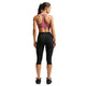 En kvinde står med ryggen til og har en Nike-sport-BH med polstring til kvinder i farven Pink Smoke/White på. Hun har desuden sorte Nike-capri-leggings med det ikoniske Swoosh-logo og Dri-FIT-teknologi samt sorte sportssko på. Hendes hår er sat op i en hestehale foran en hvid baggrund.