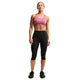 En kvinde står med ansigtet vendt fremad iført Nike Padded Sports Bra, Dame i farven Pink Smoke/White, samt sorte capri-leggings og sorte Nike-sportssko med det karakteristiske Swoosh-logo. Hendes hår er sat op, og hun holder armene afslappet ned langs siden foran en ensfarvet hvid baggrund.