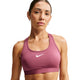 En person, der bærer Nike Padded Sports Bra, Dame i farven Pink Smoke/White, står foran en ensfarvet baggrund og ser direkte ind i kameraet. Håret er sat op, og personen bærer små ringøreringe.