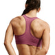 En kvinde ses bagfra iført Nike Padded Sports Bra, Dame, i farven Pink Smoke/White. Hendes hår er sat op, og hendes arme er let bøjet ved albuerne. Den polstrede sports-BH træder tydeligt frem mod den ensfarvede hvide baggrund og tilbyder både stil og støtte.