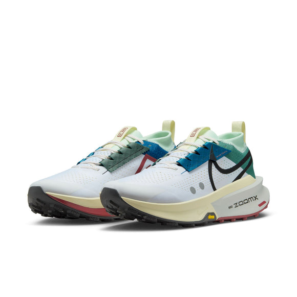 Et par Nike Zegama 2 Herre-sneakers i White/Black-Bicoastal-Court Blue med blå og røde detaljer, sorte Swoosh-logoer, lysegrønne detaljer, tykke ZoomX-skumsåler og Vibram Megagrip-ydersåler, der giver et godt greb i terrænet.