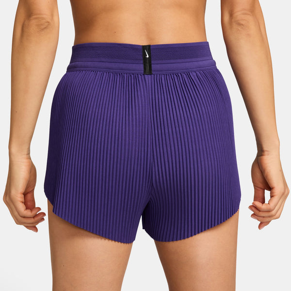 En person ses fra taljen og ned iført Nike Aeroswift 3" løbeshorts til kvinder i Court Purple/White, med høj talje, plisseret, struktureret stof og et lille sort mærke midt på linningen.