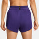 En person ses fra taljen og ned iført Nike Aeroswift 3" løbeshorts til kvinder i Court Purple/White, med høj talje, plisseret, struktureret stof og et lille sort mærke midt på linningen.