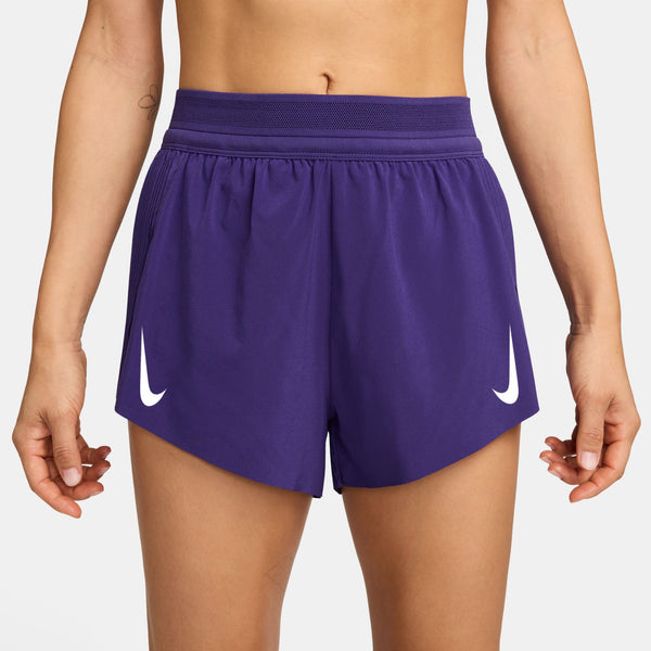 En person ses fra taljen til midt på låret iført Nike Aeroswift 3" løbeshorts til kvinder i Court Purple/White med hvide logoer og en elastisk linning med Dri-FIT ADV-teknologi. Armene er afslappede i siden.