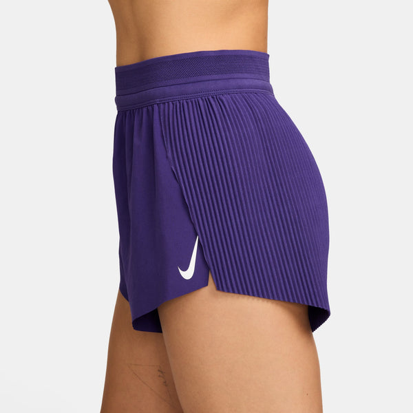 En kvinde er iført Nike Aeroswift 3" løbeshorts i Court Purple/White, der er fremstillet af bæredygtige materialer og har et hvidt Nike-logo på venstre side. Billedet er taget fra siden mod en almindelig hvid baggrund.